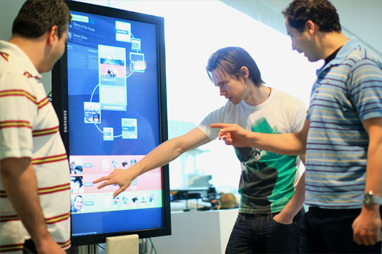 The Glass Infrastructure , a network of 30 touch screens placed throughout the facility, was created to make the Media Lab's website tangible. The screens recognize employees and visitors by their identification tags. The system then guides individuals around the building and provides background information on the people and projects they might encounter. Here, Boris Kizelshteyn, Greg Elliott, and Polychronis Ypodimatopoulos interact with one of the screens.
