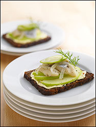 Smorbord: Hearty bread sliced thin and spread with butter is piled high with apple slices, pickled herring, and cucumber, then garnished with a sprig of dill.