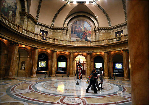 Women walk through Memorial Hall at the State House. The hall is ringed with murals.