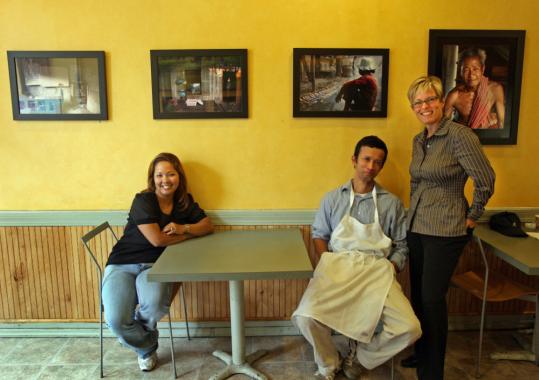 Nancy Carbonaro (right) with her photos of Cambodia at the Wellesley bakery run by siblings Sambo Rattanavong (left) and Mara Nuon; their family's story includes fleeing the Khmer Rouge.