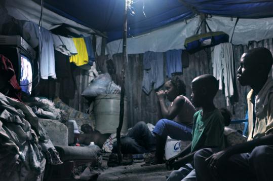 A family gathered in a makeshift tent in Port-au-Prince to watch TV. Many Haitians living in camps after last year’s earthquake say their misery is exploited by foreign aid groups.